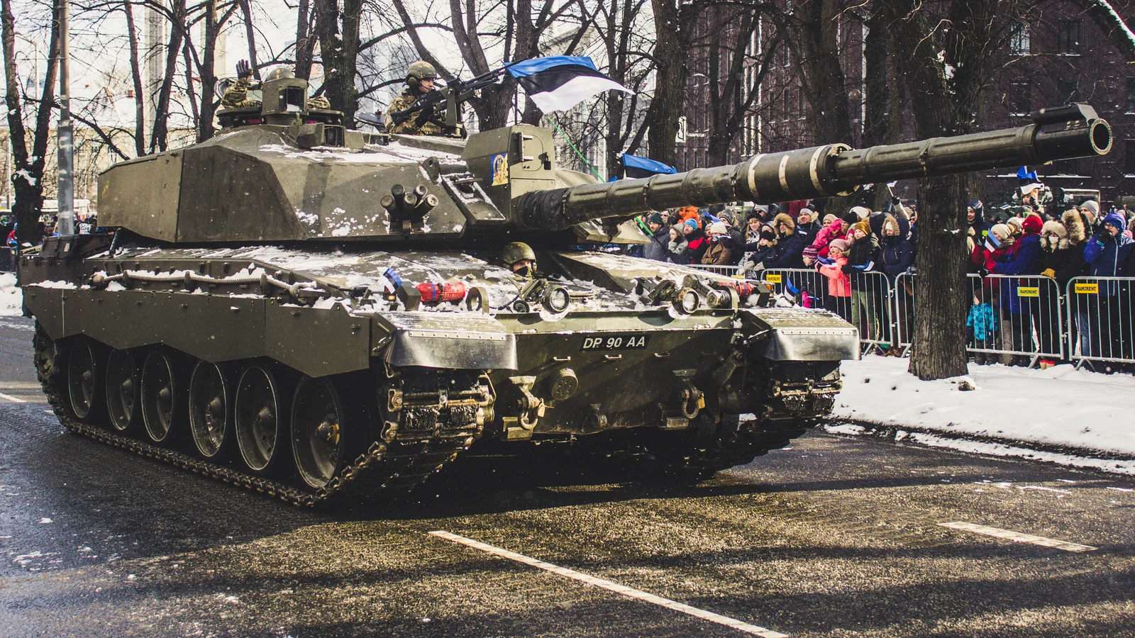 Carro de combate británico Challenger 2 desfilando por una calle nevada de Estonia, con público y banderas estonias al fondo