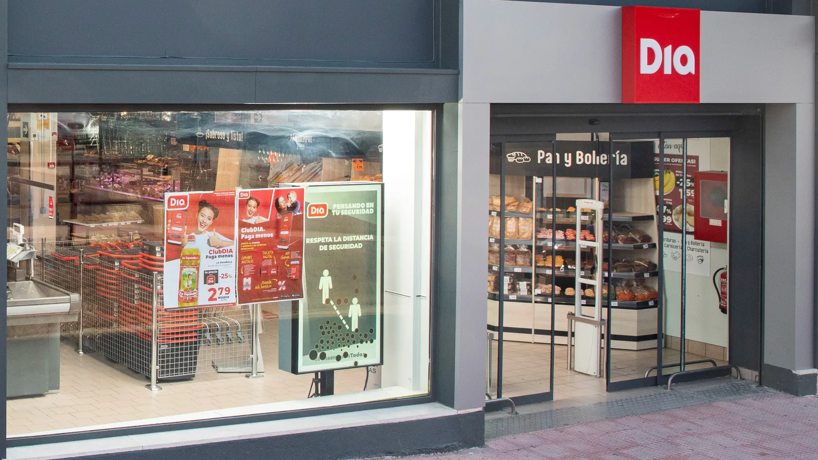 Fachada de un supermercado Dia con su rótulo rojo y la sección de pan y bollería visible tras el escaparate.