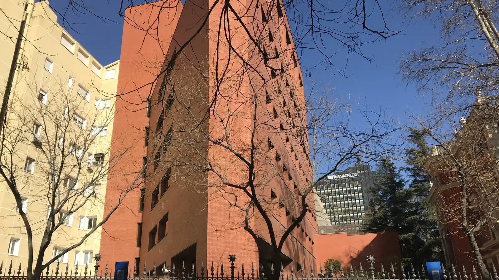 Edificio de ladrillo rojo de Bankinter en Madrid visto desde la calle, con árboles sin hojas delante, una verja en primer plano y el cielo azul de fondo.