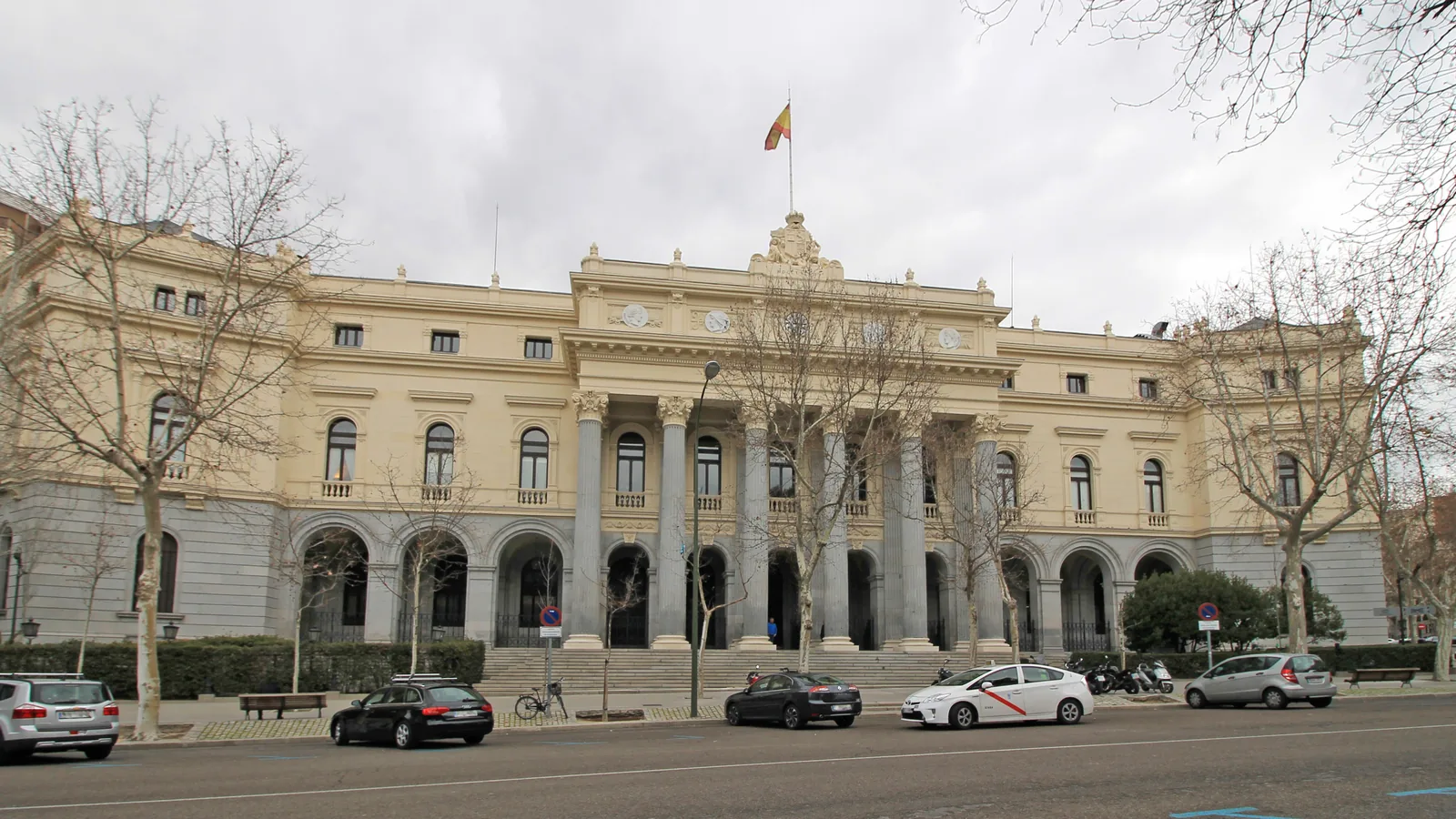 Fachada neoclásica del Palacio de la Bolsa de Madrid, con columnas, escalinata y bandera de España, vista desde la calle con coches y taxis aparcados delante.