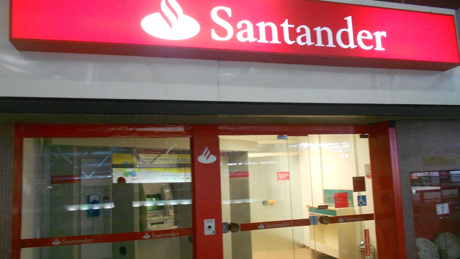 Rótulo rojo iluminado de Santander sobre la entrada acristalada de una oficina bancaria en un aeropuerto de Brasil.