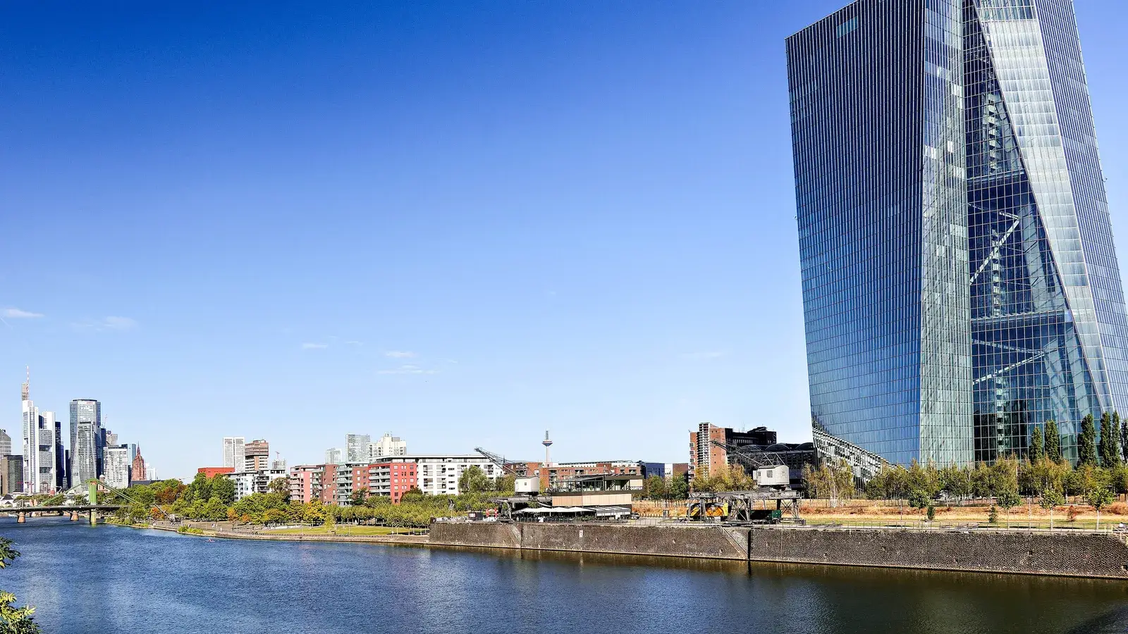 Torre de cristal de la sede del Banco Central Europeo junto al río Meno, con el skyline de Fráncfort al fondo y cielo azul.