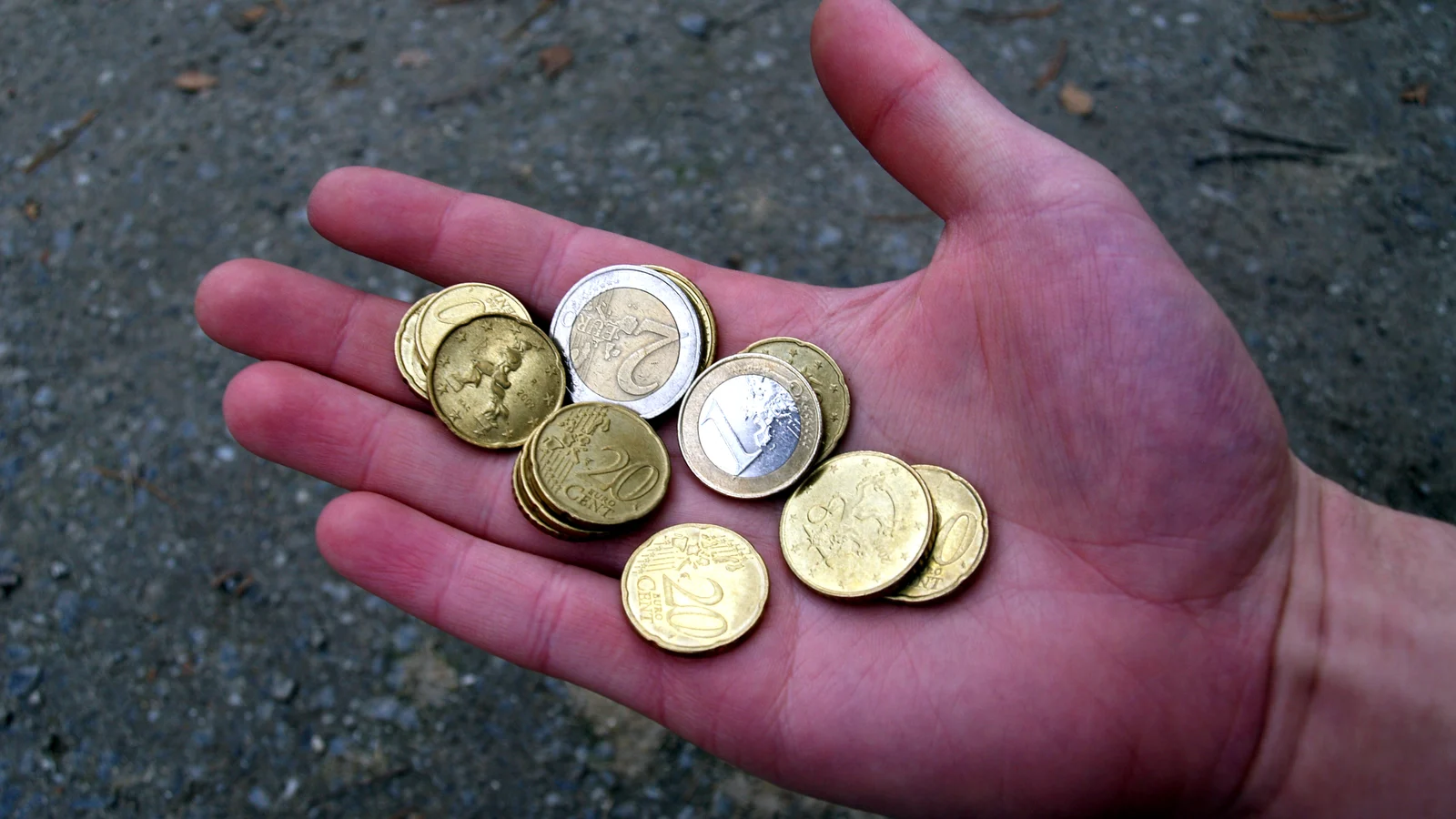 Puñado de monedas de euro, entre ellas de 2 euros, 1 euro y 20 céntimos, sobre la palma abierta de una mano, con el suelo de asfalto de fondo.