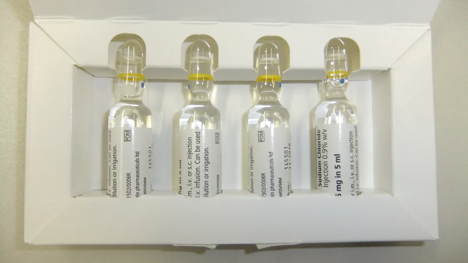 Cuatro ampollas de vidrio con tapón de anillo amarillo, colocadas en una caja de cartón blanca abierta sobre una mesa gris.